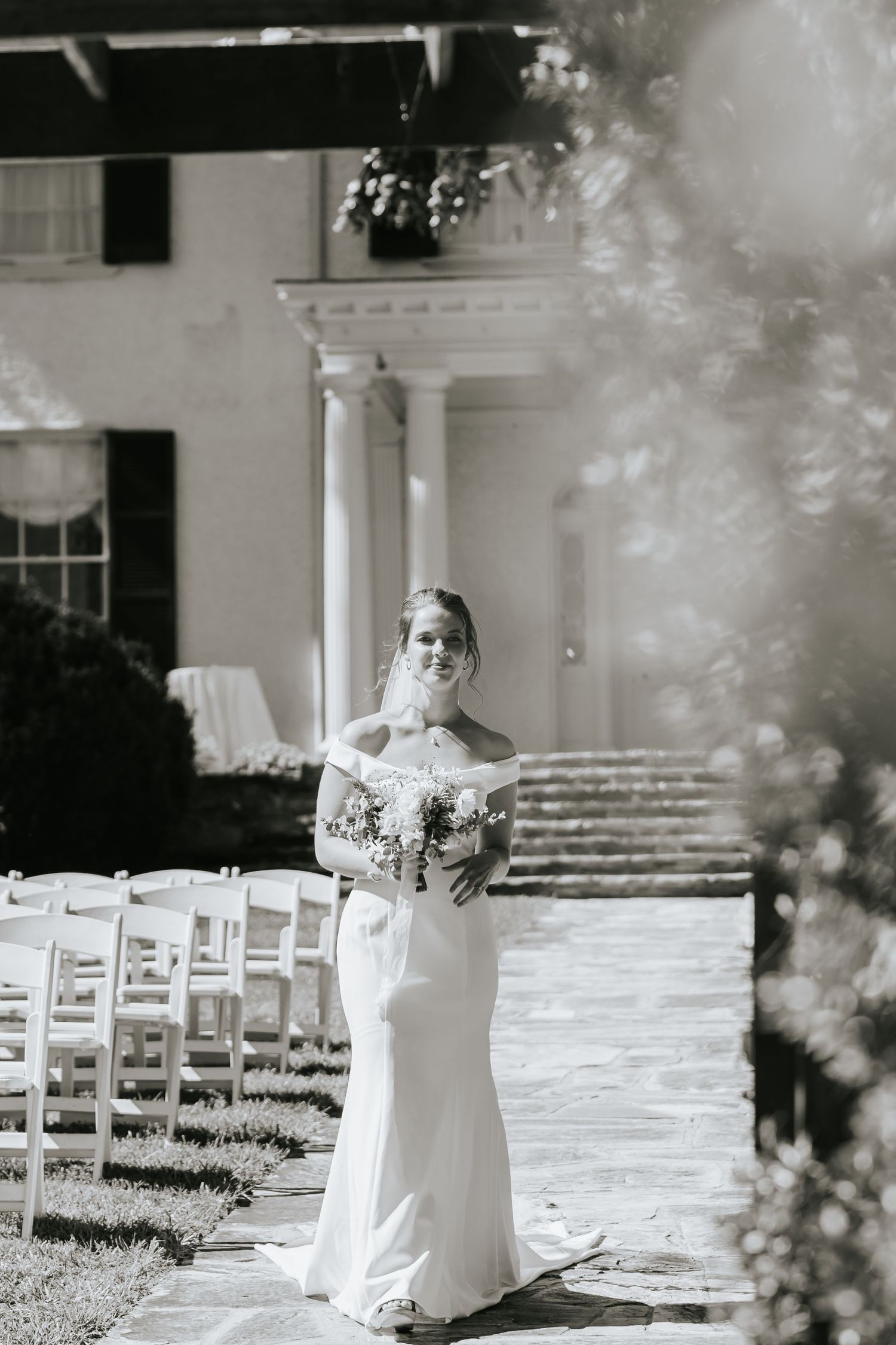 A figure in a white wedding gown stands on a stone path with white chairs and columns in the background.