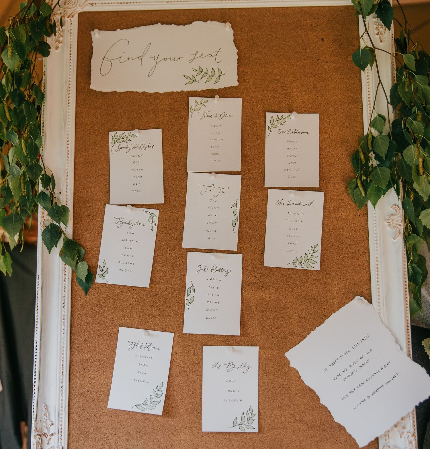 A rustic cork board displays elegant wedding seating arrangements with white cards and green vine decorations.
