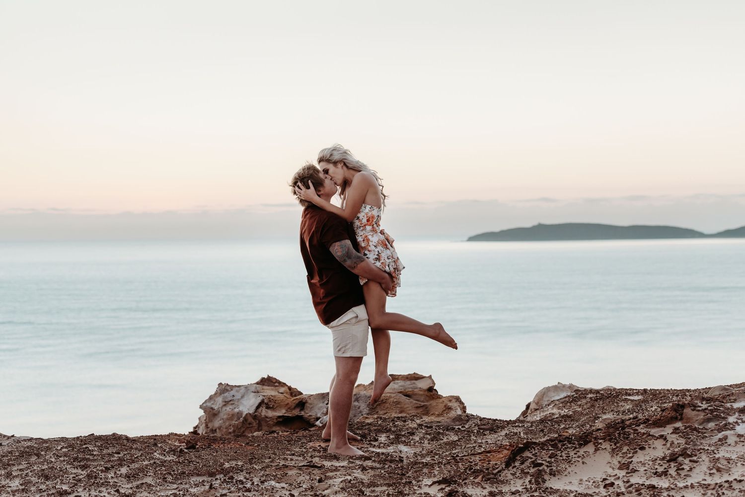 Couple embracing on coastal cliff edge during sunset with ocean view.