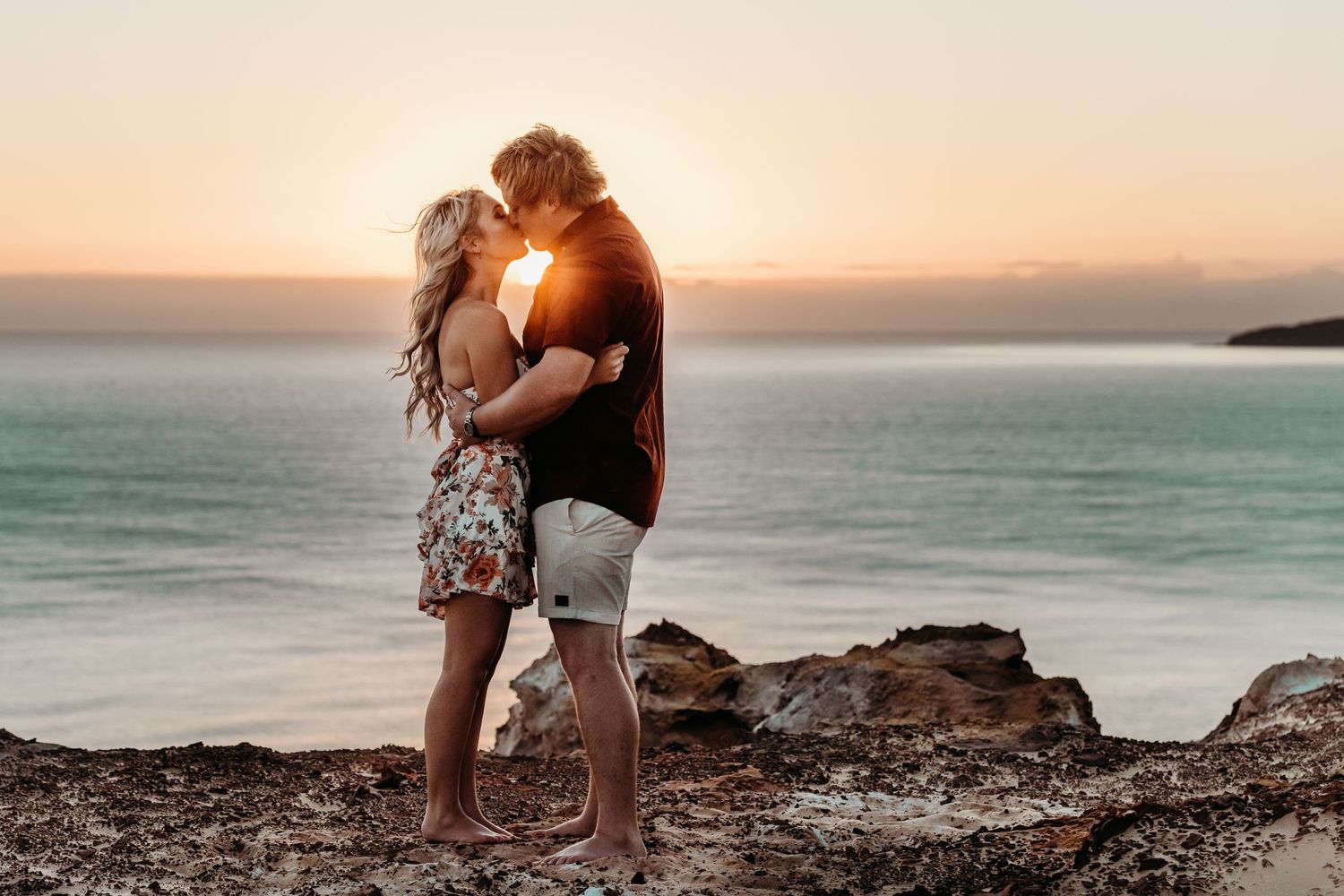 A couple embraces on rocky coastal cliffs during golden hour sunset.