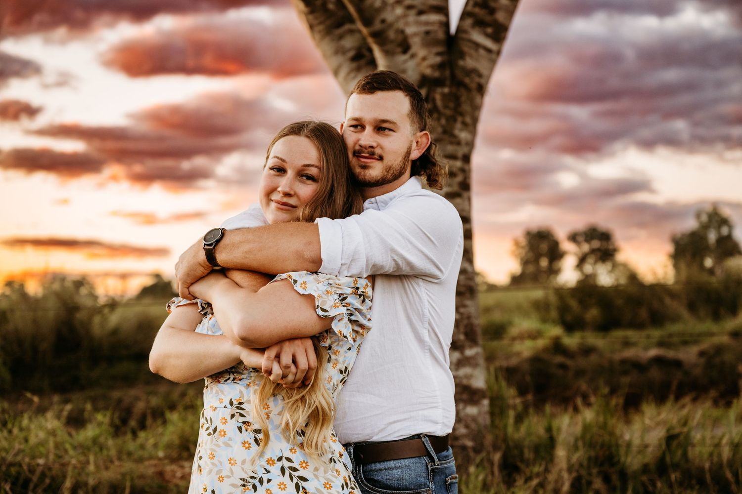 A tender embrace captured in a field during golden hour with dramatic sunset clouds.