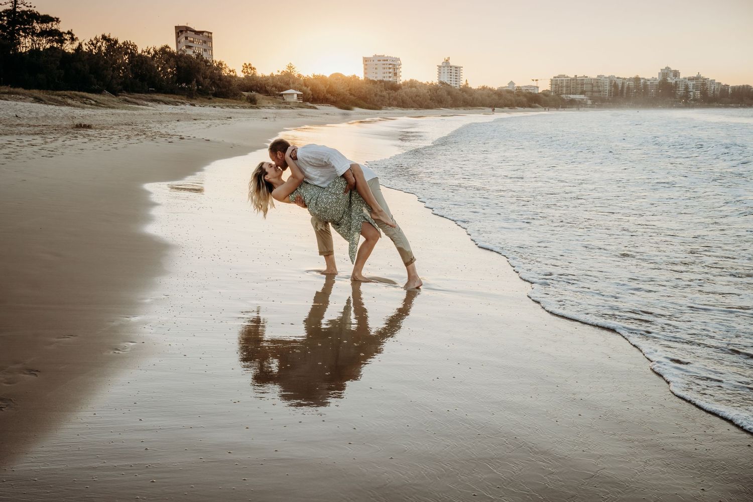 A couple plays in the ocean waves at sunset with their reflection in the wet sand.