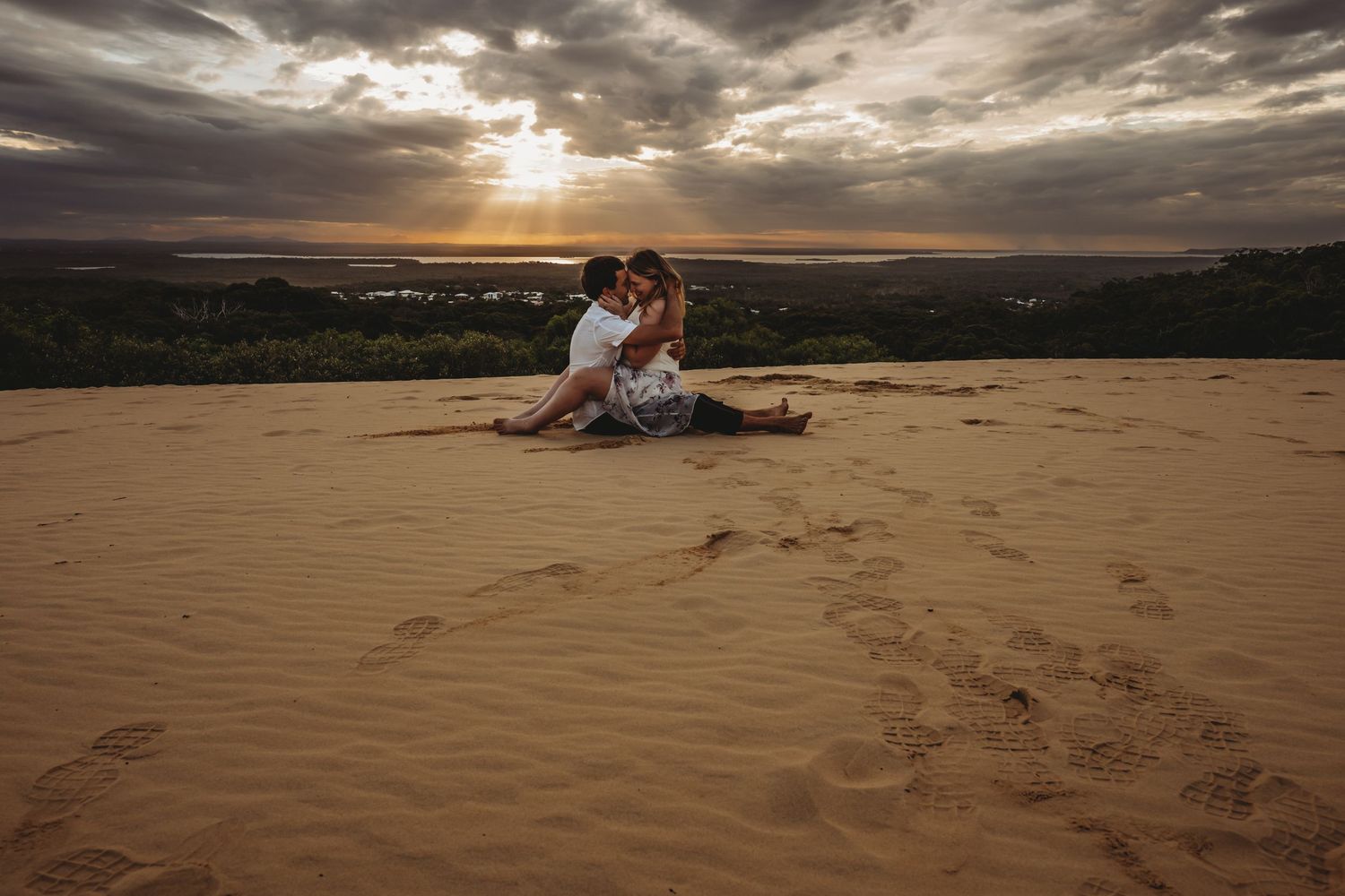 A couple sits together on sandy dunes during a dramatic sunset with clouds.