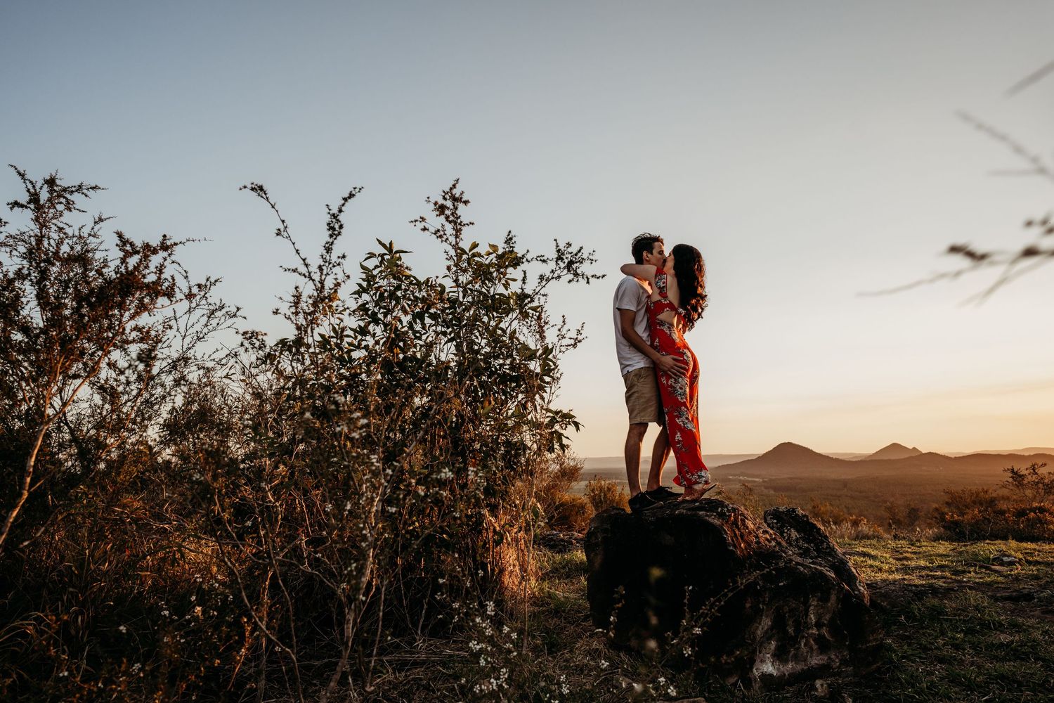 A couple stands close together on a rock formation at sunset in a desert landscape.