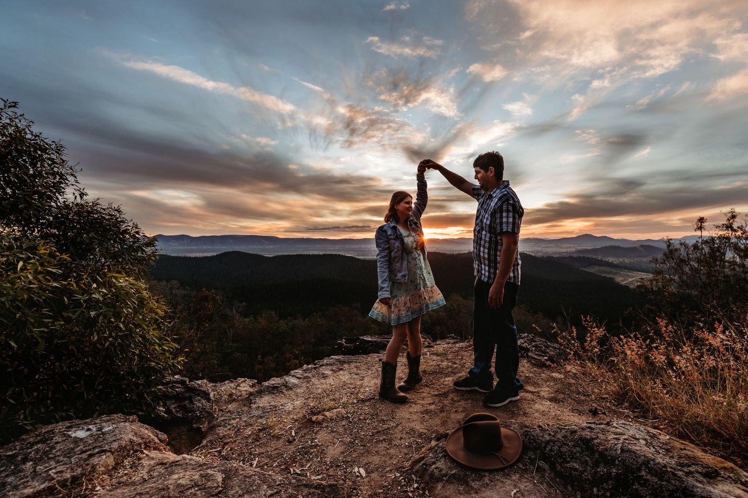 Two silhouettes stand on a mountain peak against a dramatic sunset sky with clouds and distant mountains.