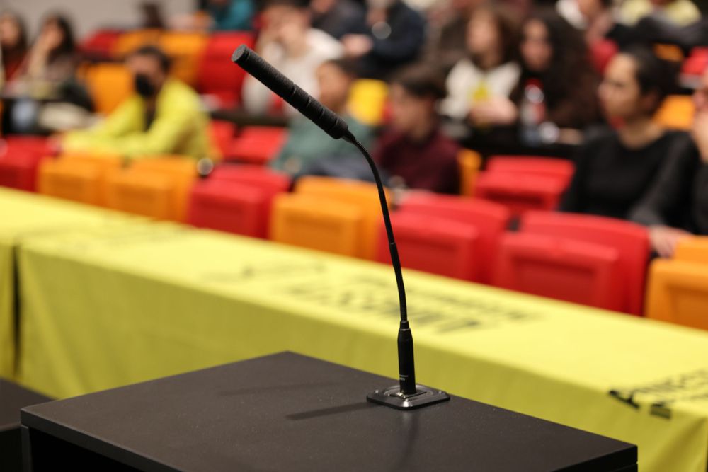 Empty microphone stand on podium with colorful chairs in background at conference venue.