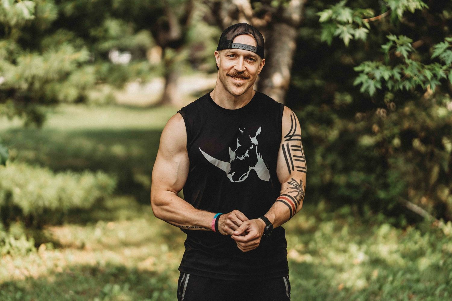 Athletic person in black tank top poses outdoors with muscular arms and tribal tattoo.