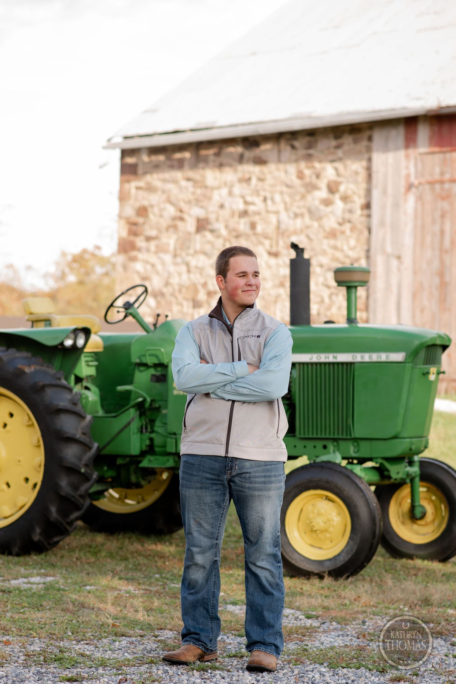 Classic John Deere tractors parked beside stone barn with person in casual attire standing.