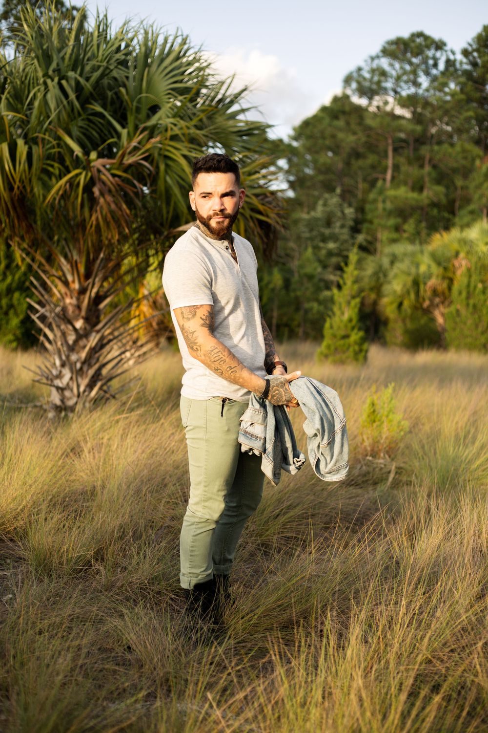 Person in casual attire holds jacket while posing near palm trees in natural setting.