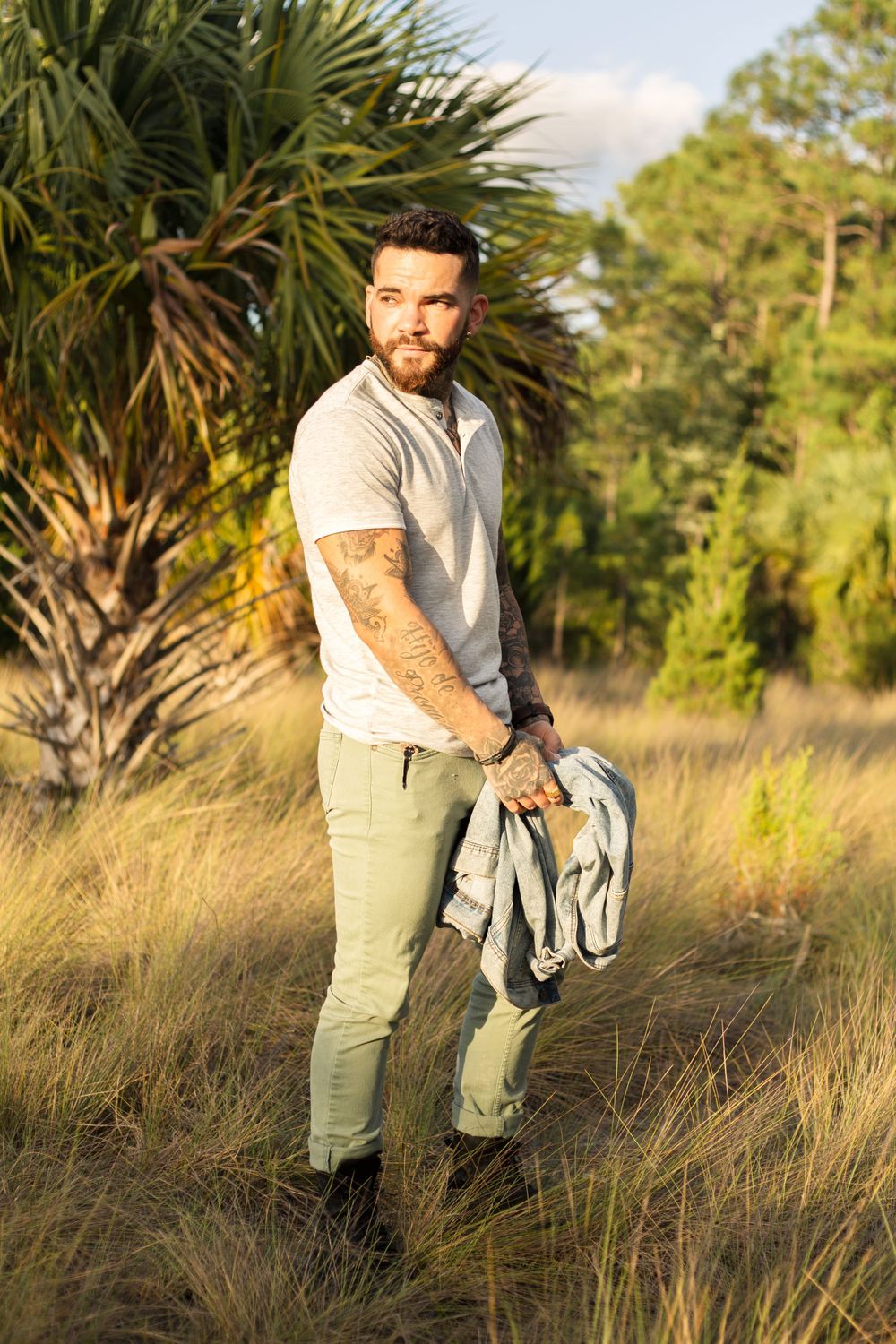 Casual dressed person with tattoos poses while holding jacket in coastal landscape.