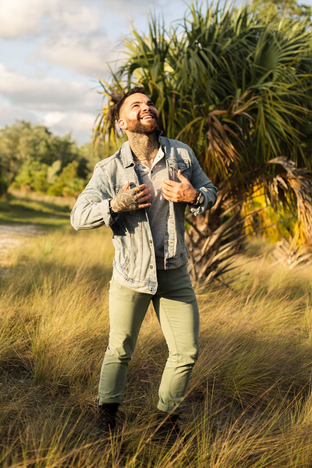 Person in denim jacket smiles while standing in golden grass with palm trees.