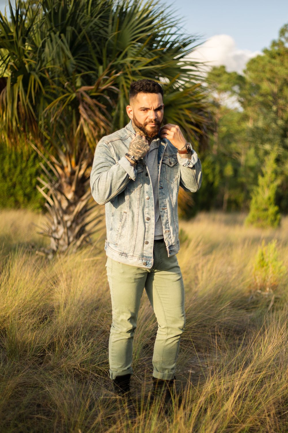 Person in light denim jacket standing in tall grass with palm trees in background during golden hour.