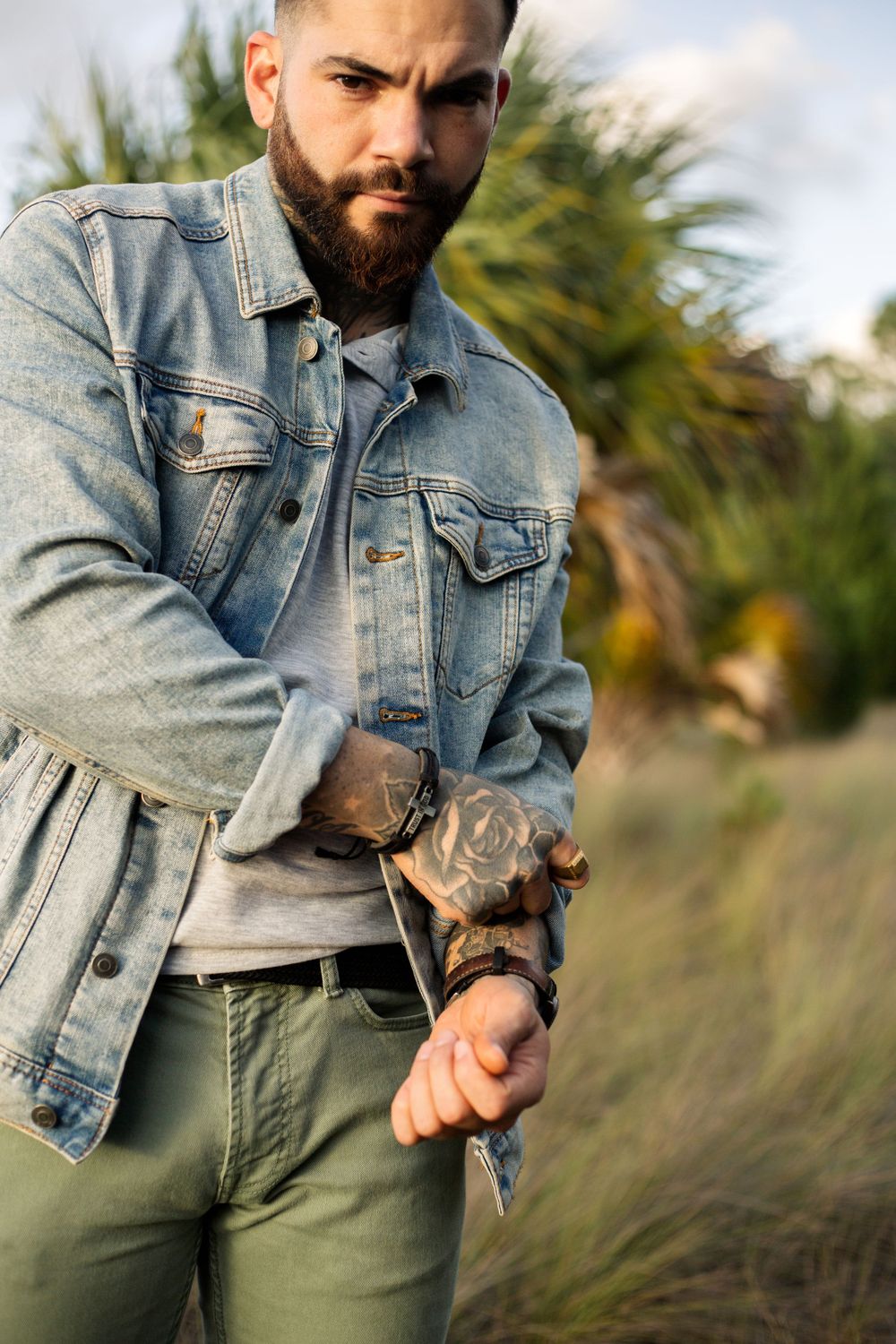 Person wearing denim jacket and olive pants showing detailed arm tattoos in outdoor setting.