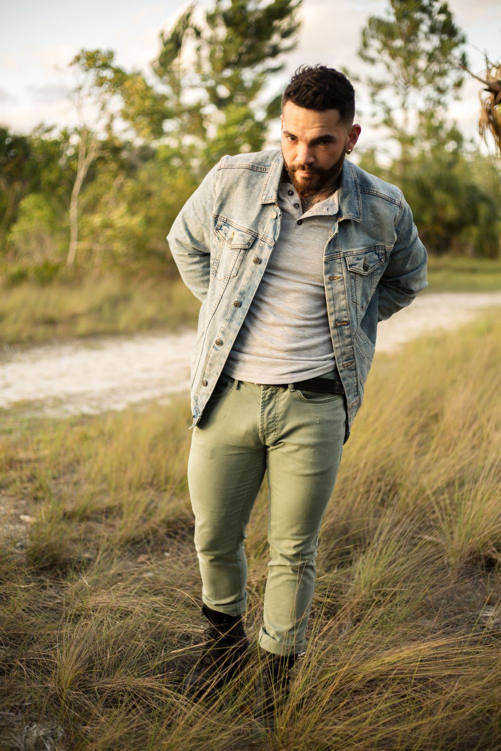 A person in casual wear including denim jacket and olive pants poses in tall grass during golden hour.