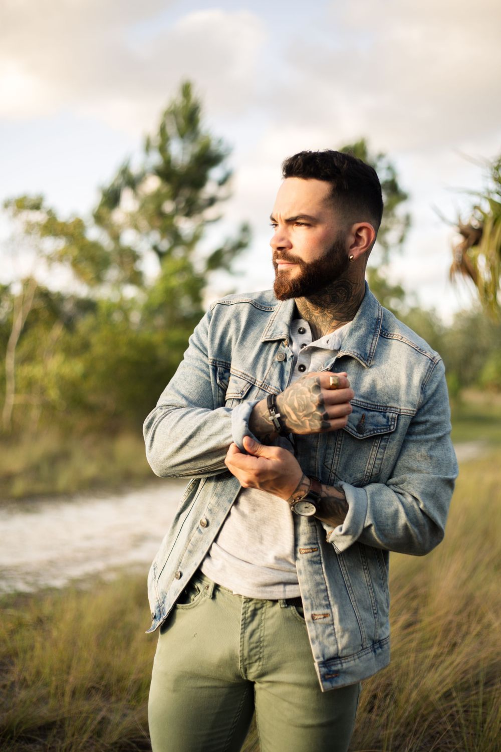 Person adjusting denim jacket sleeve while standing in natural outdoor setting.