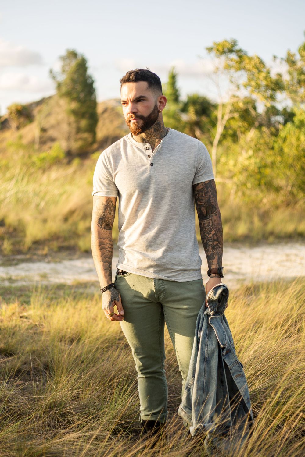 Person in grey henley shirt and olive pants holding denim jacket in grassy field.