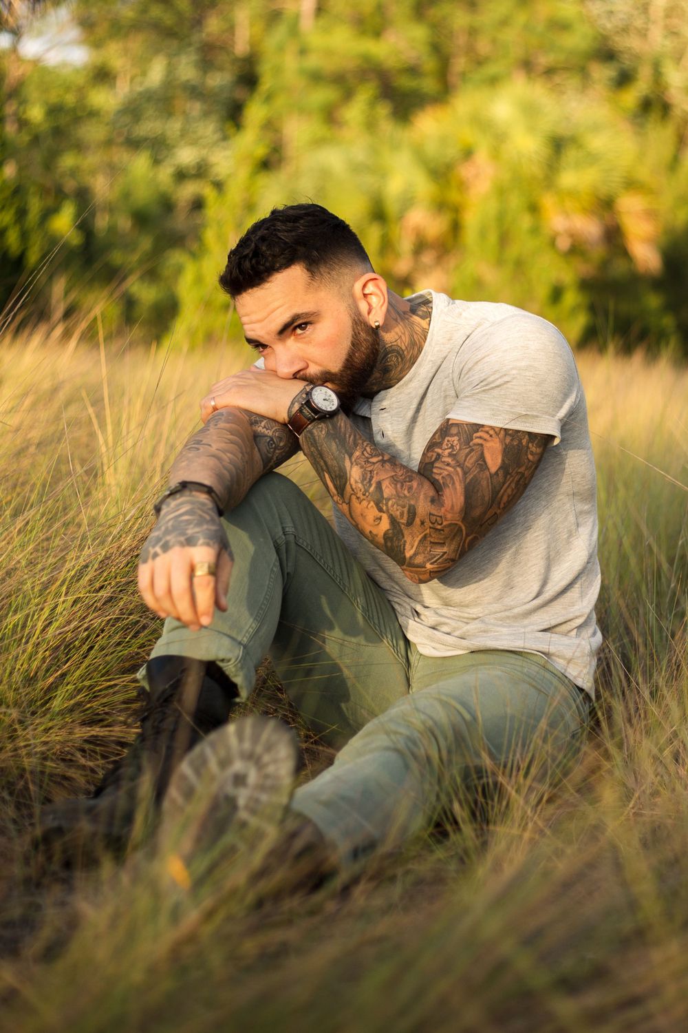 Person with tattoos sitting in tall grass during sunset wearing casual attire.