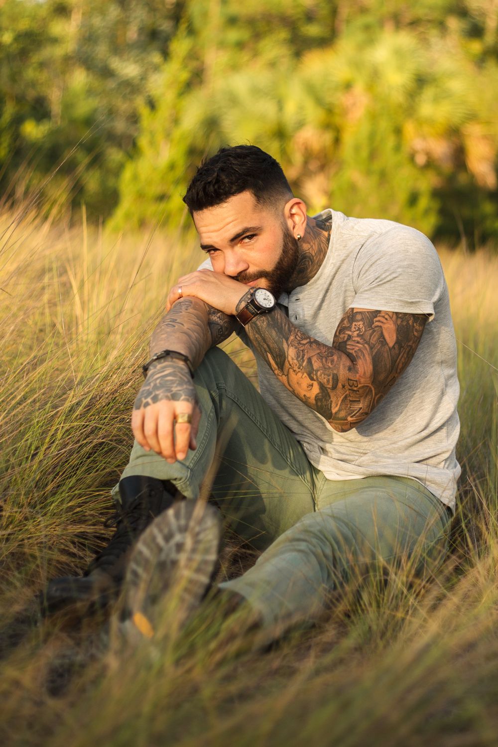 A tattooed person in casual attire sits contemplatively in a field of tall grass.