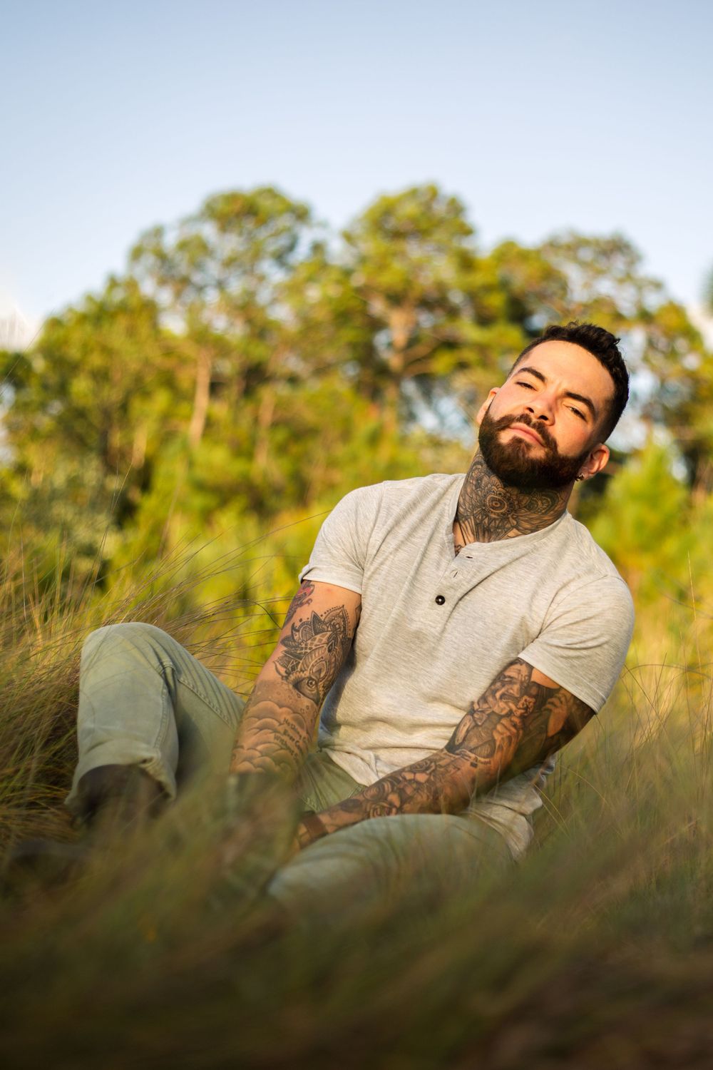 A bearded person with tattoos relaxes in tall grass at sunset.
