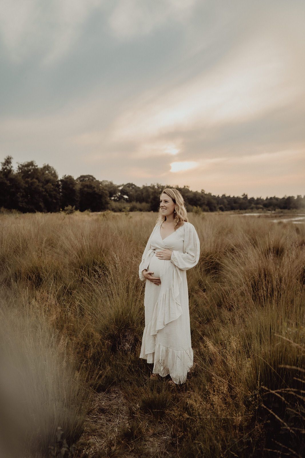 A dreamy maternity photoshoot in a golden field at sunset features a flowing white lace dress and natural surroundings.