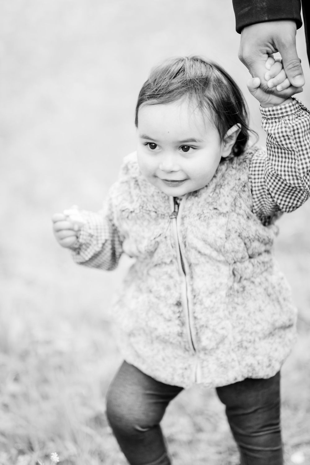A black and white photo of a toddler in a plush vest holding someone's hand while walking.