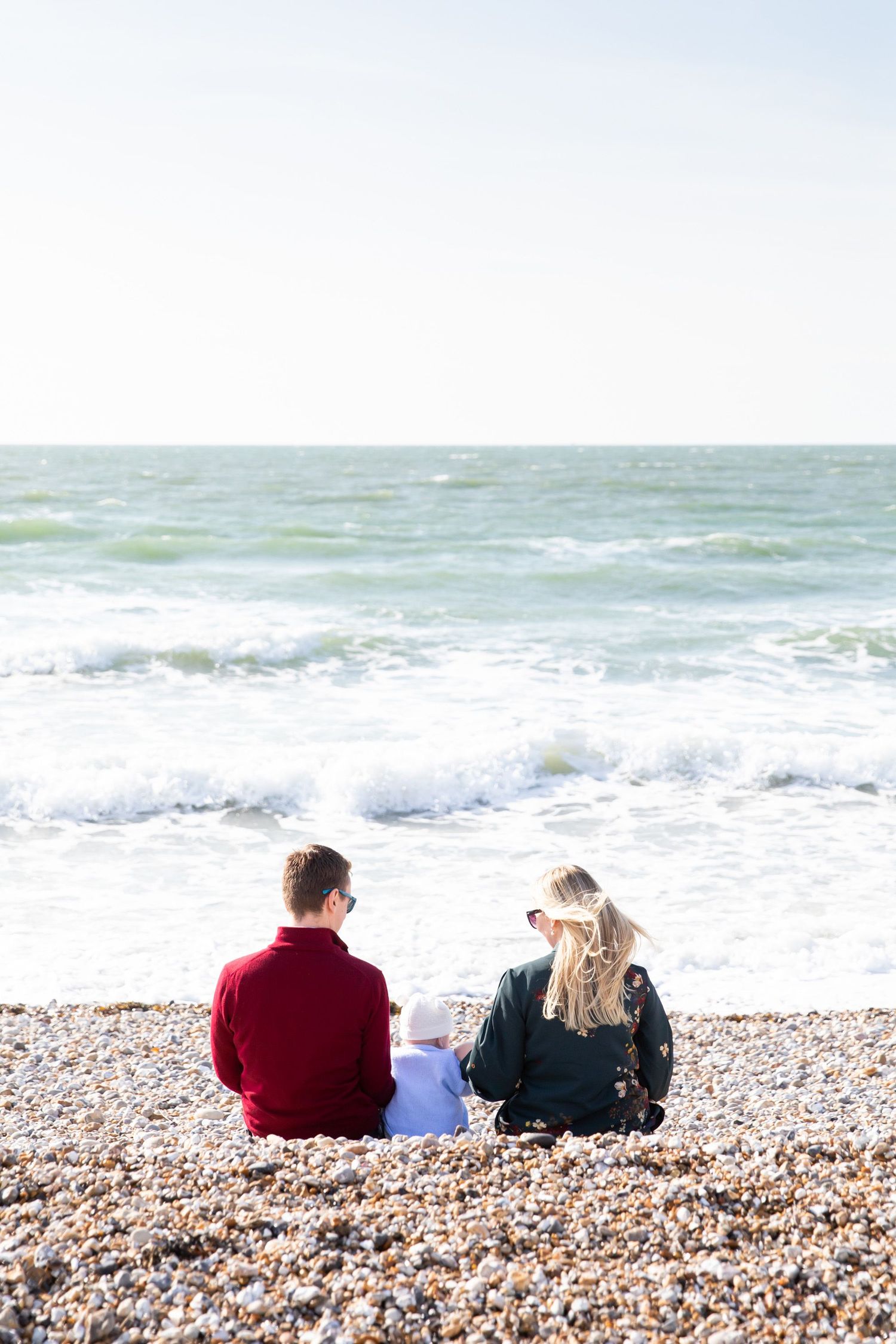 Family sits on pebble beach watching waves crash on shore.