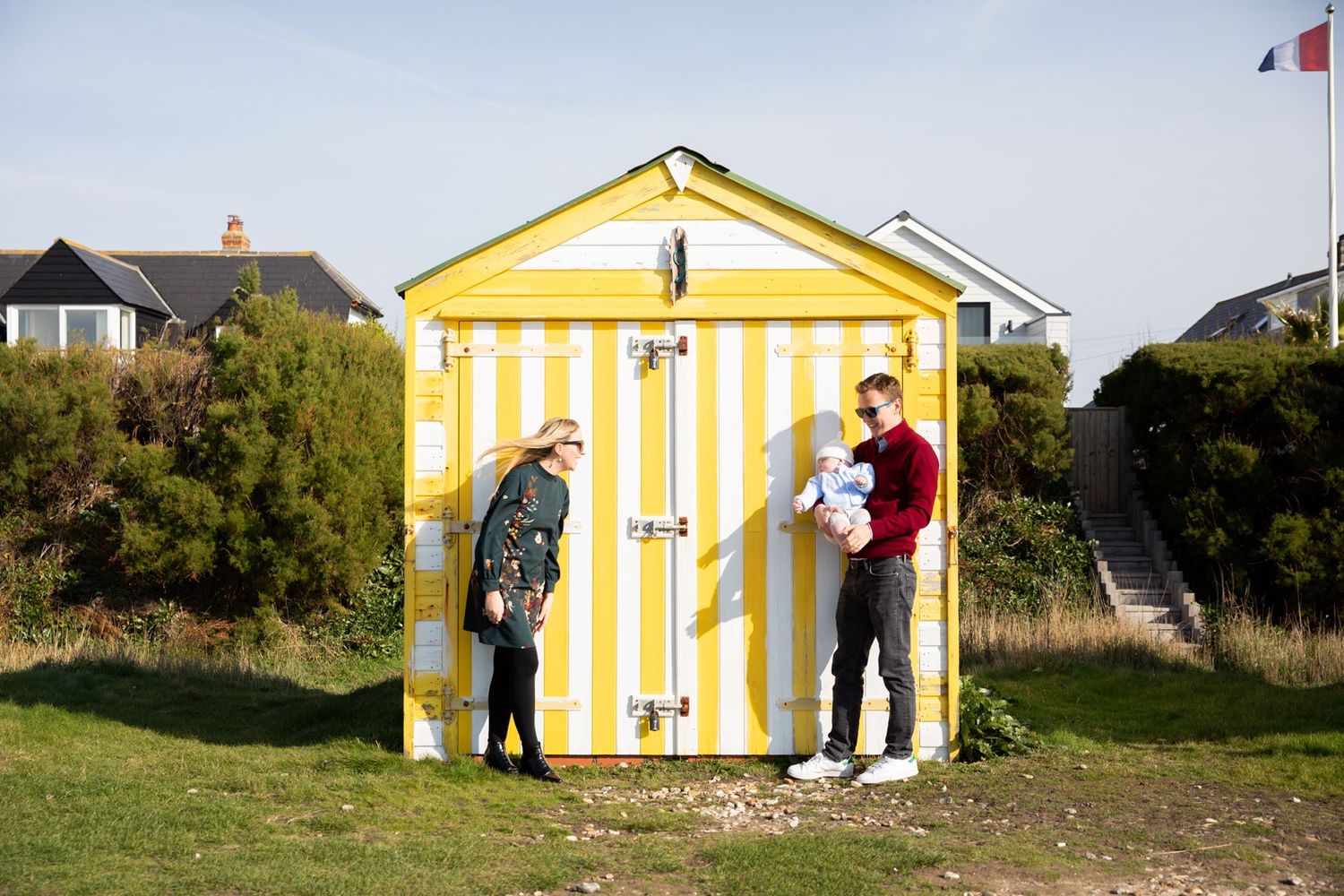 People stand beside yellow and white striped beach hut on grassy area.