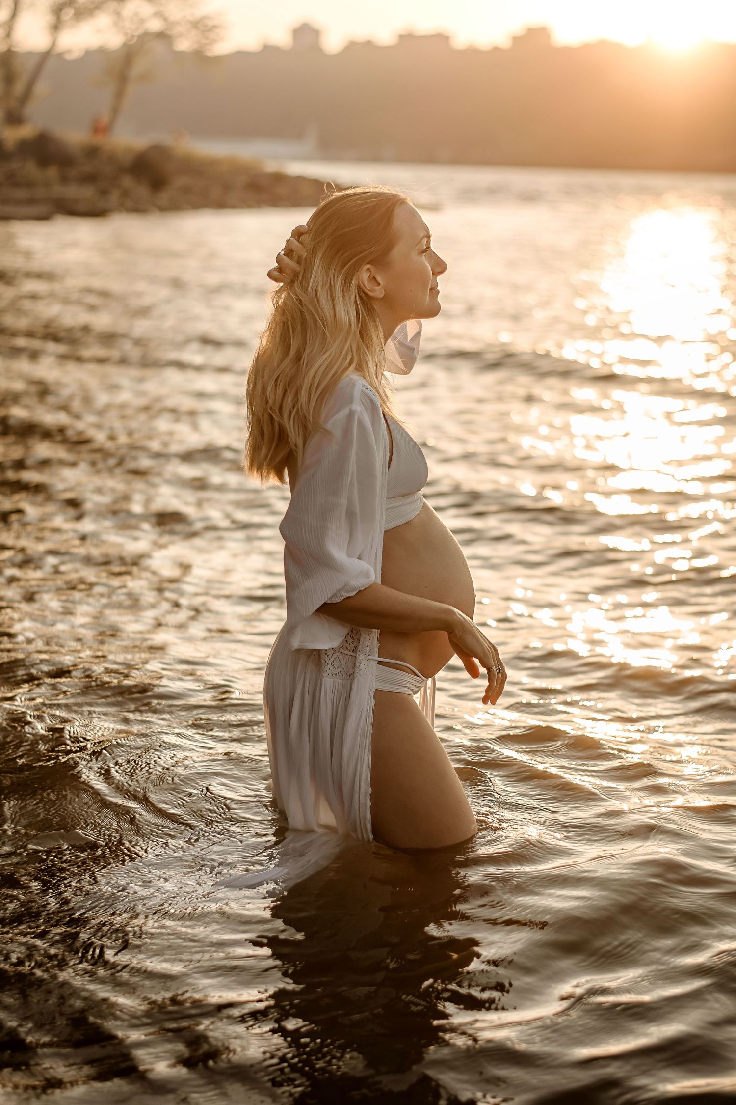 A pregnant woman stands in glowing water at sunset wearing a flowing white cover-up.