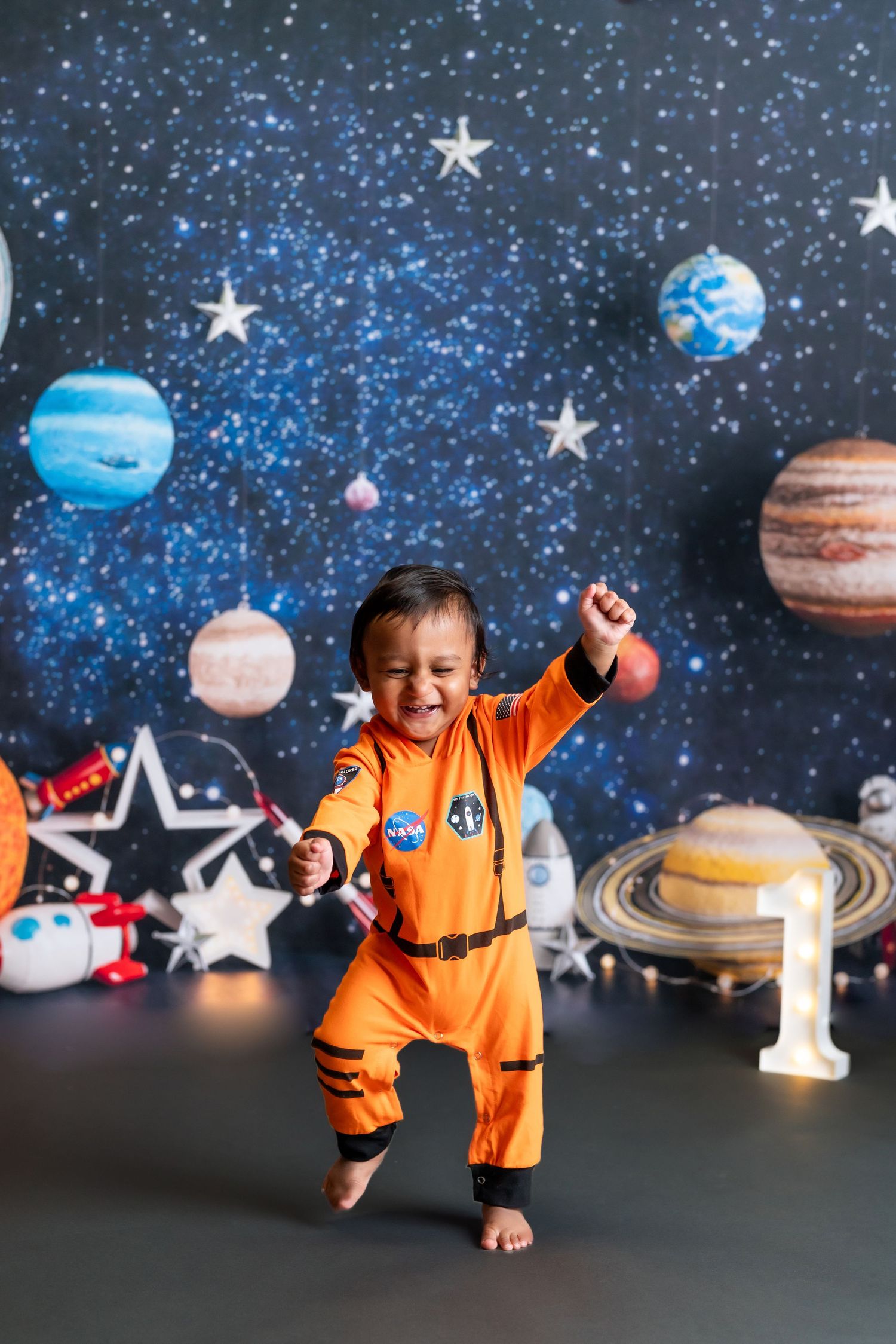 Baby in NASA-style orange jumpsuit jumping against space-themed backdrop.