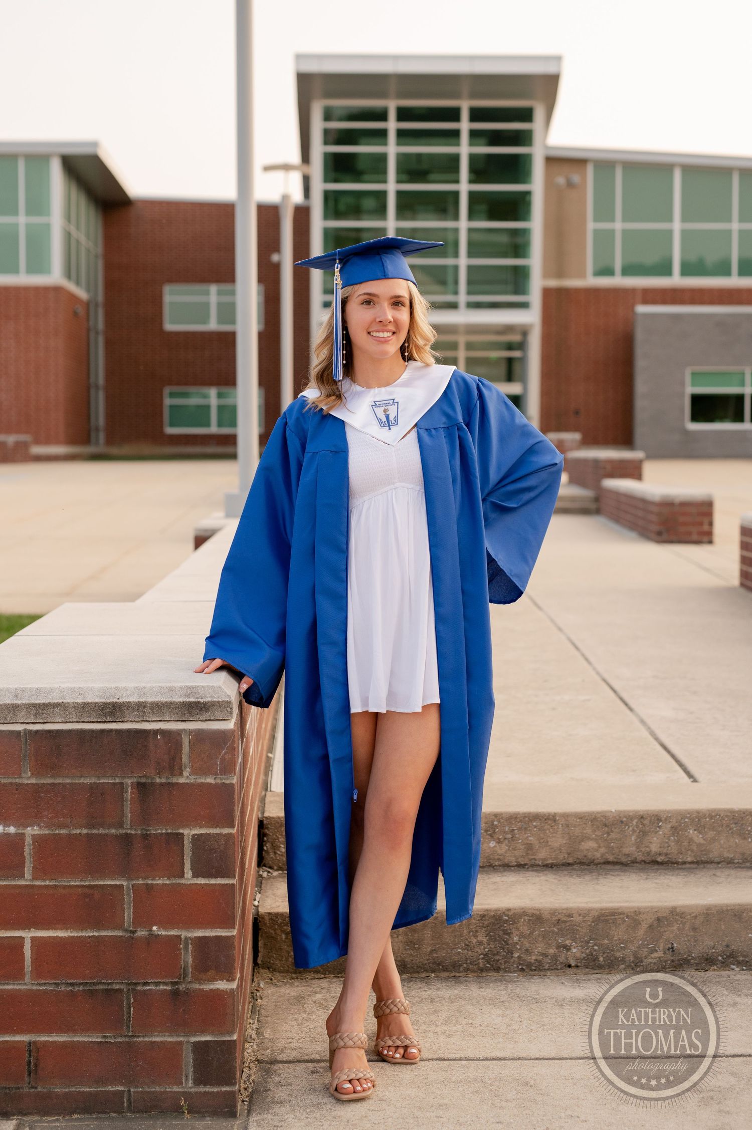 Graduate wearing a blue commencement gown poses confidently on campus steps.