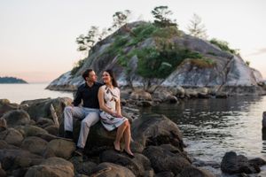 A couple sits together on coastal rocks with a dramatic island backdrop at sunset.
