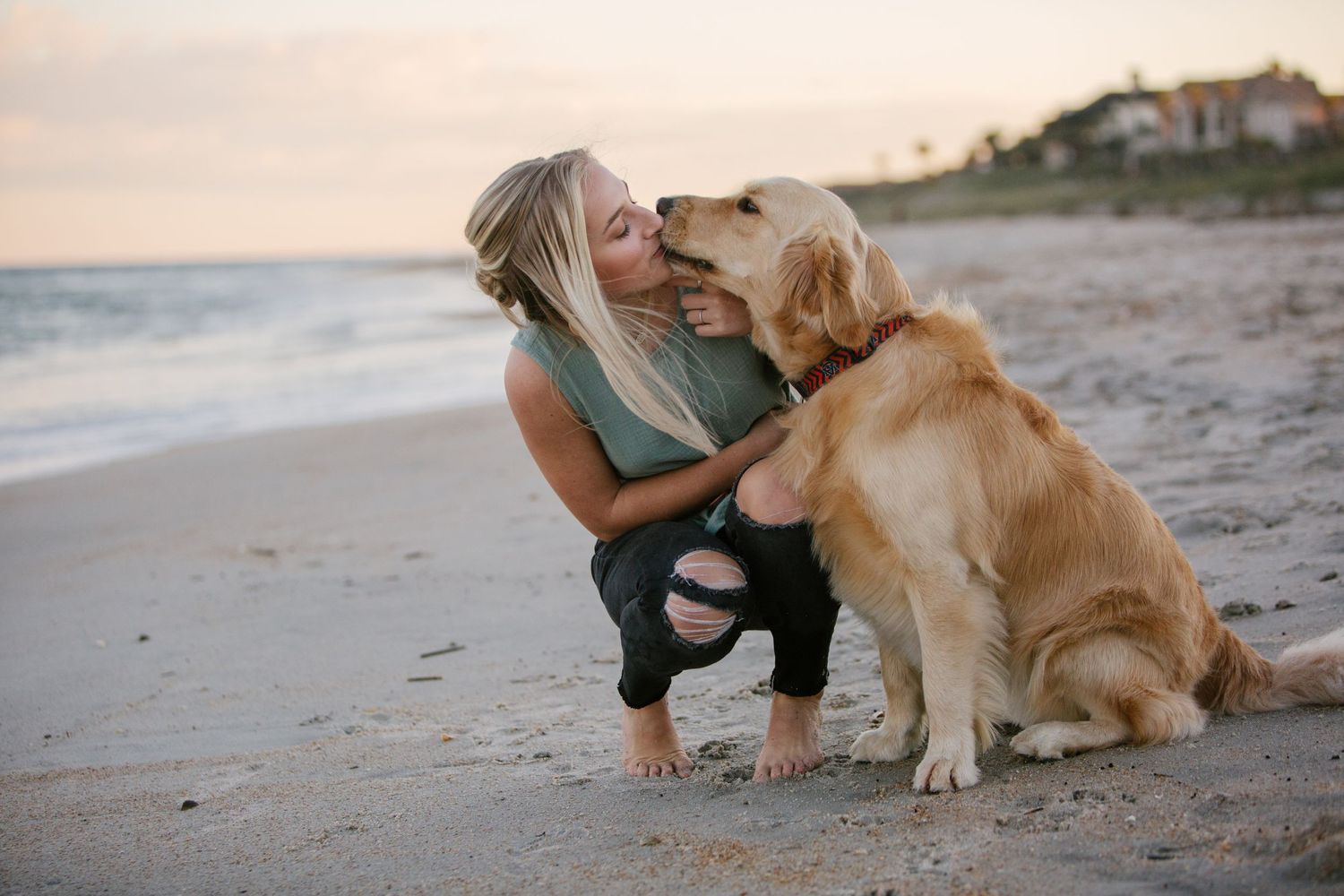 Woman in teal top playing with golden retriever on beach at sunset.