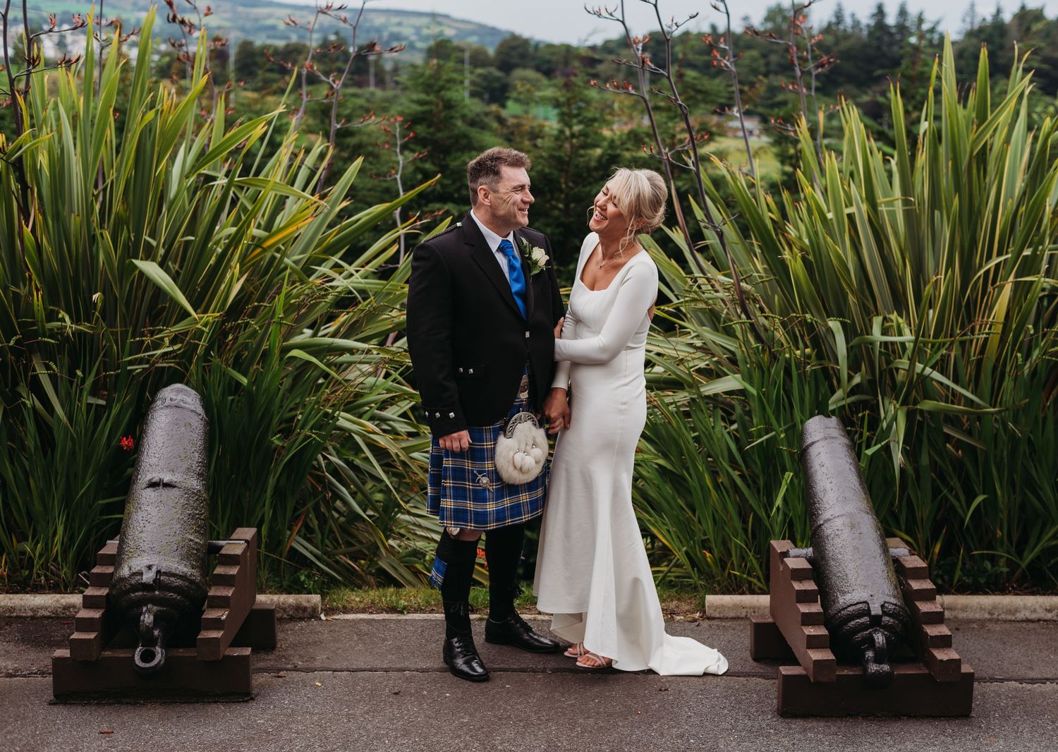 A happy couple who are laughing and holding hands. They are standing in-between two canons and greenery.