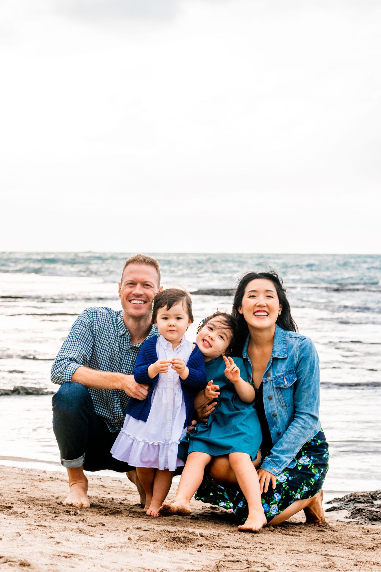 Family of four posing together on beach with ocean waves in background.