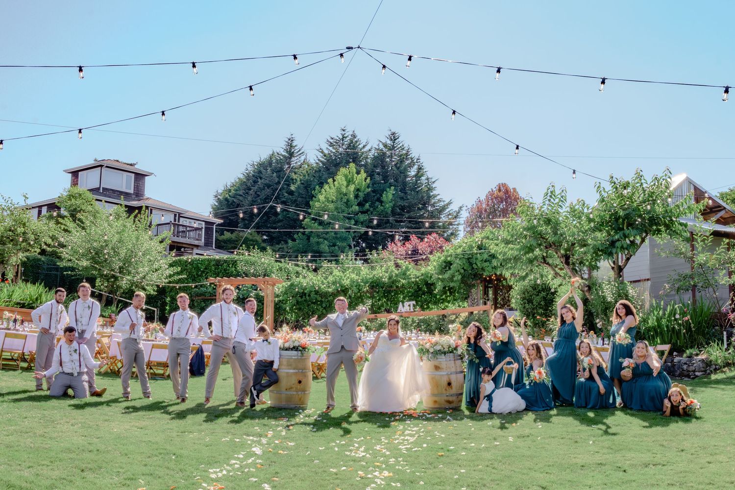 A wedding party poses together in a garden setting during a sunny outdoor celebration.