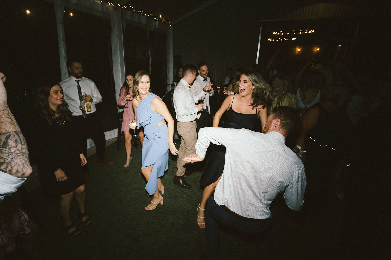 Wedding guests dancing enthusiastically on a dark dance floor under twinkling lights at a reception celebration.