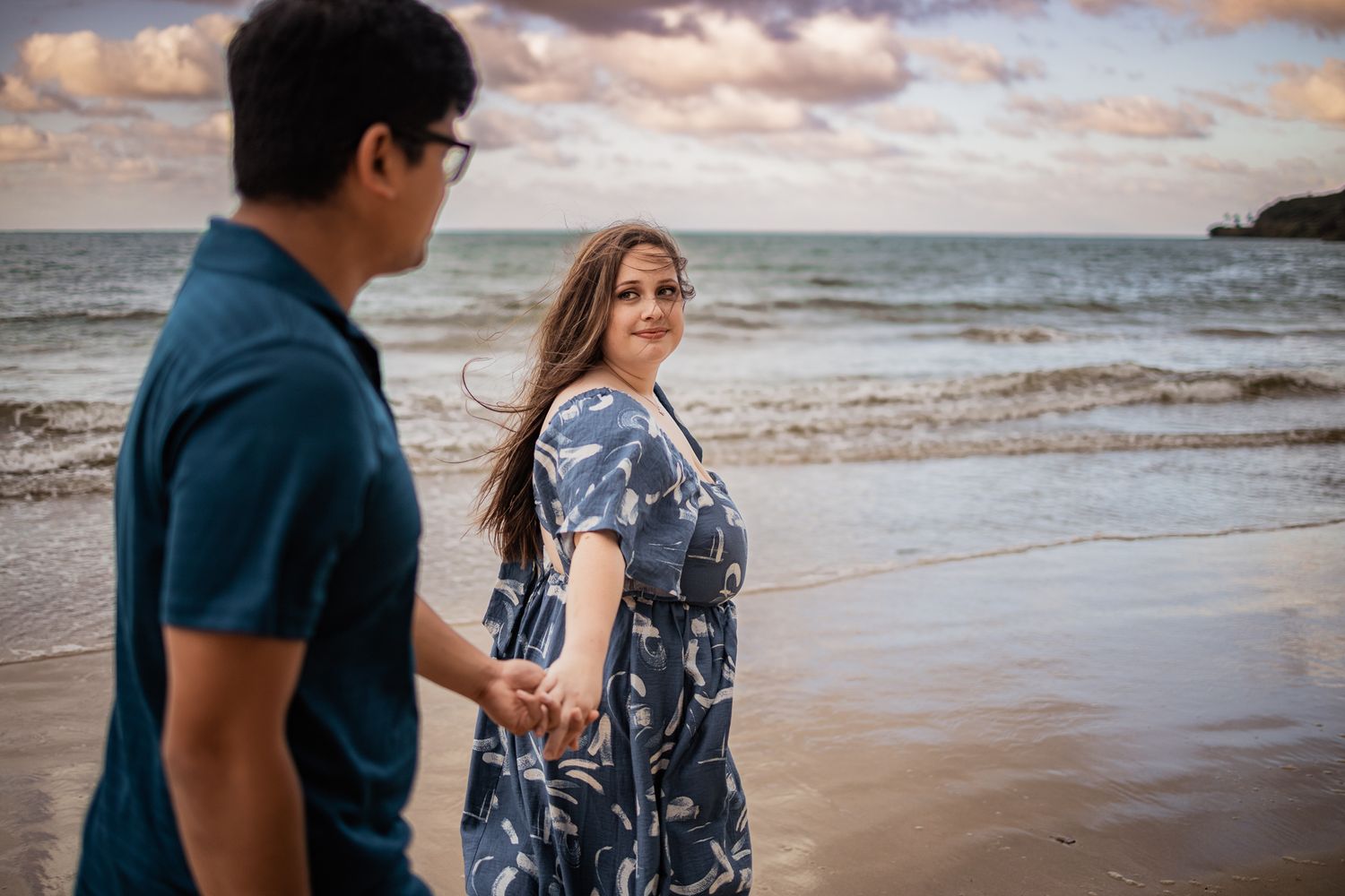 Couple walking along a scenic beach at sunset with clouds reflecting on the ocean.