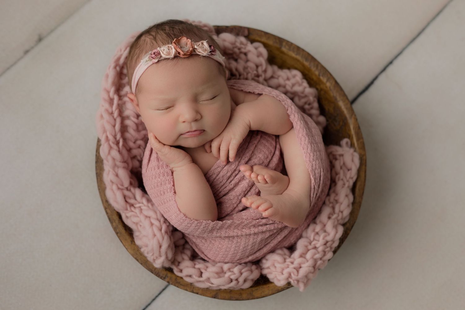 Perfectly swaddled baby girl in pink and placed in a cute wooden bowl prop for her studio newborn photoshoot as she sleeps away.