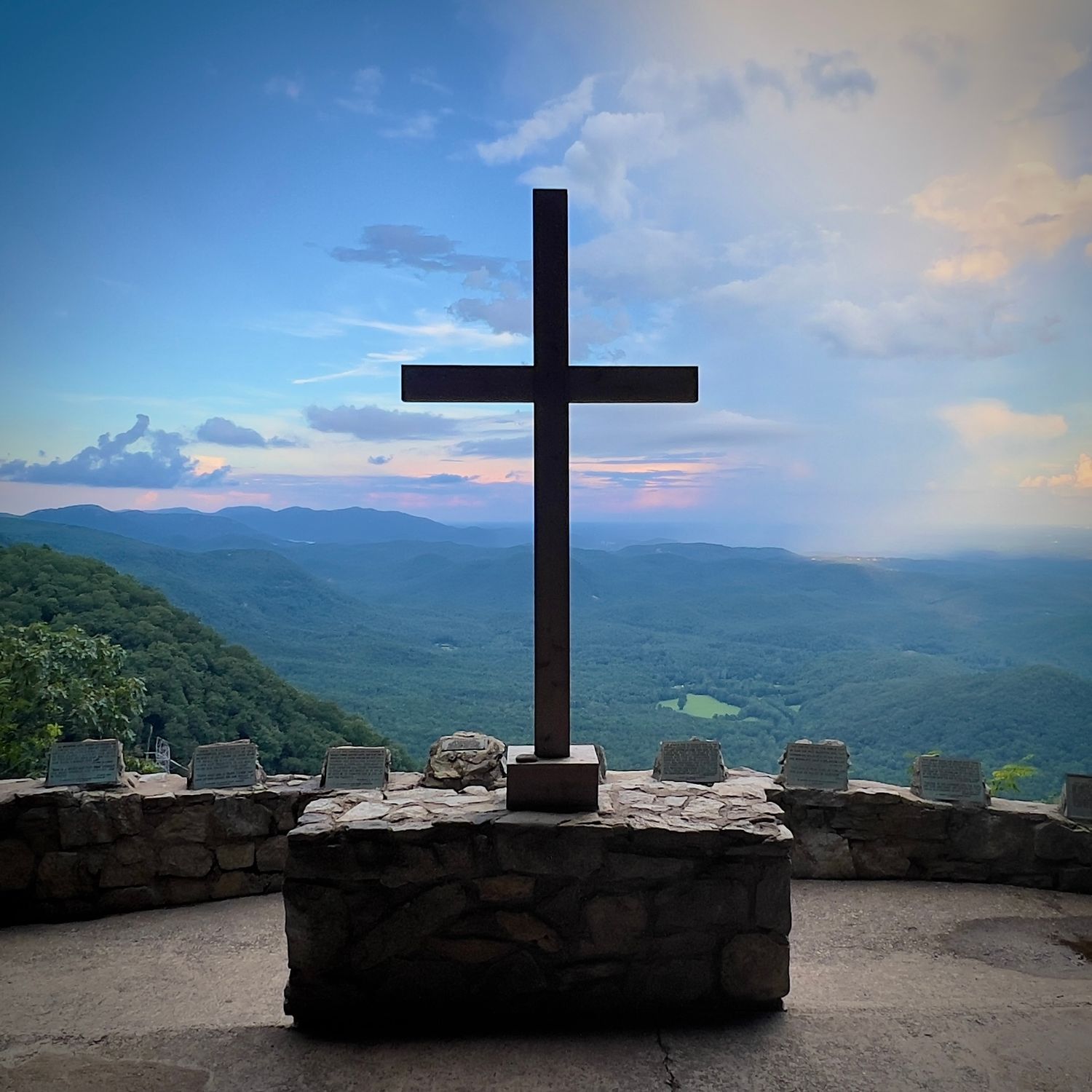 Large Christian cross monument on stone base overlooking mountain vista at sunset.