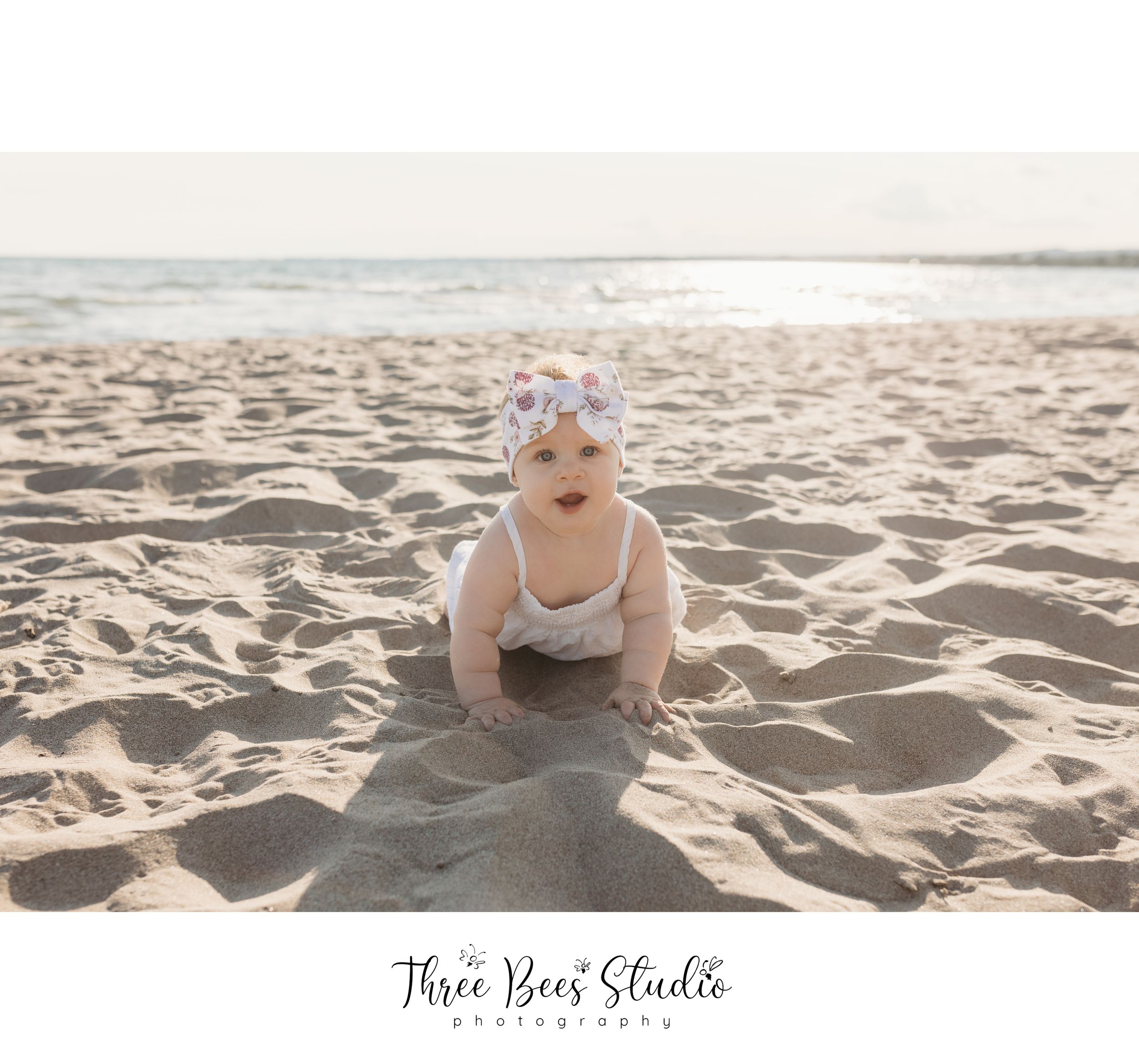 Baby crawling on sandy beach wearing white outfit and floral headband.