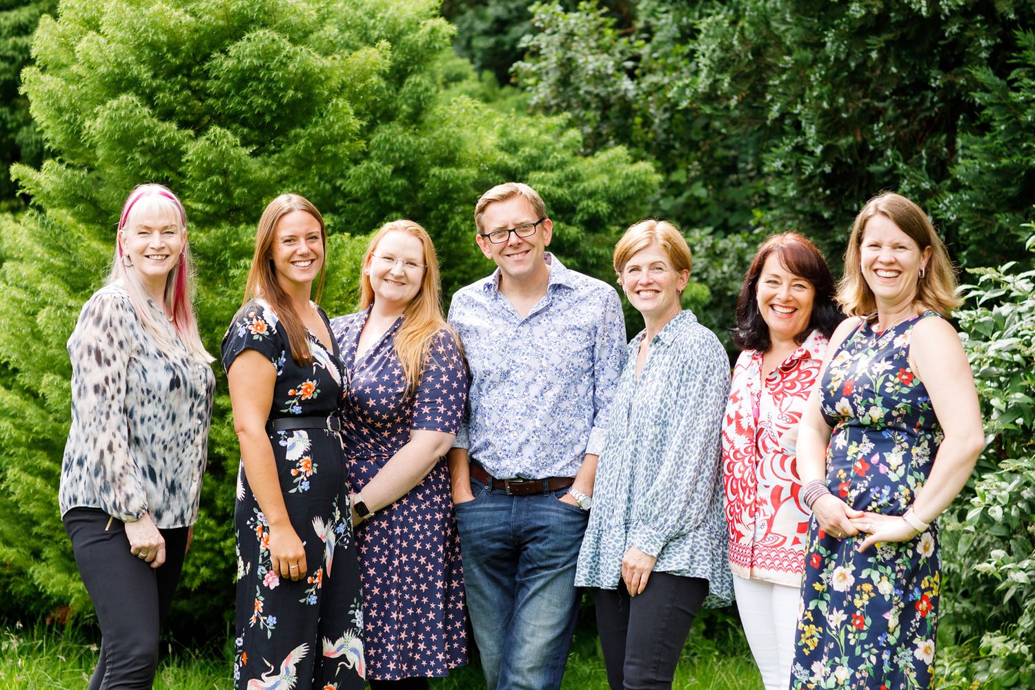 Team photo of seven people in floral prints standing together in a lush garden setting during business branding photoshoot.