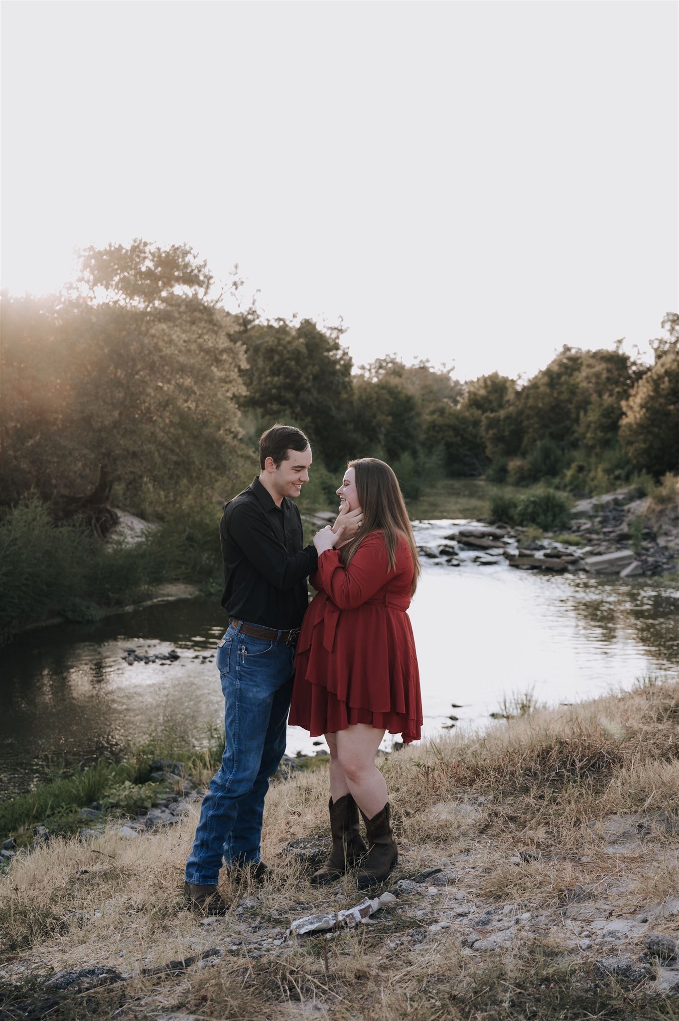 A romantic couple poses together on a hillside overlooking a stream at dusk.
