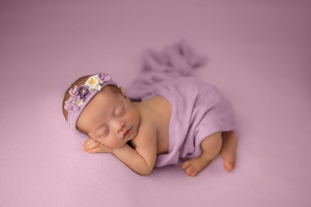 Sleeping newborn in purple wrap wearing floral headband on pink backdrop.