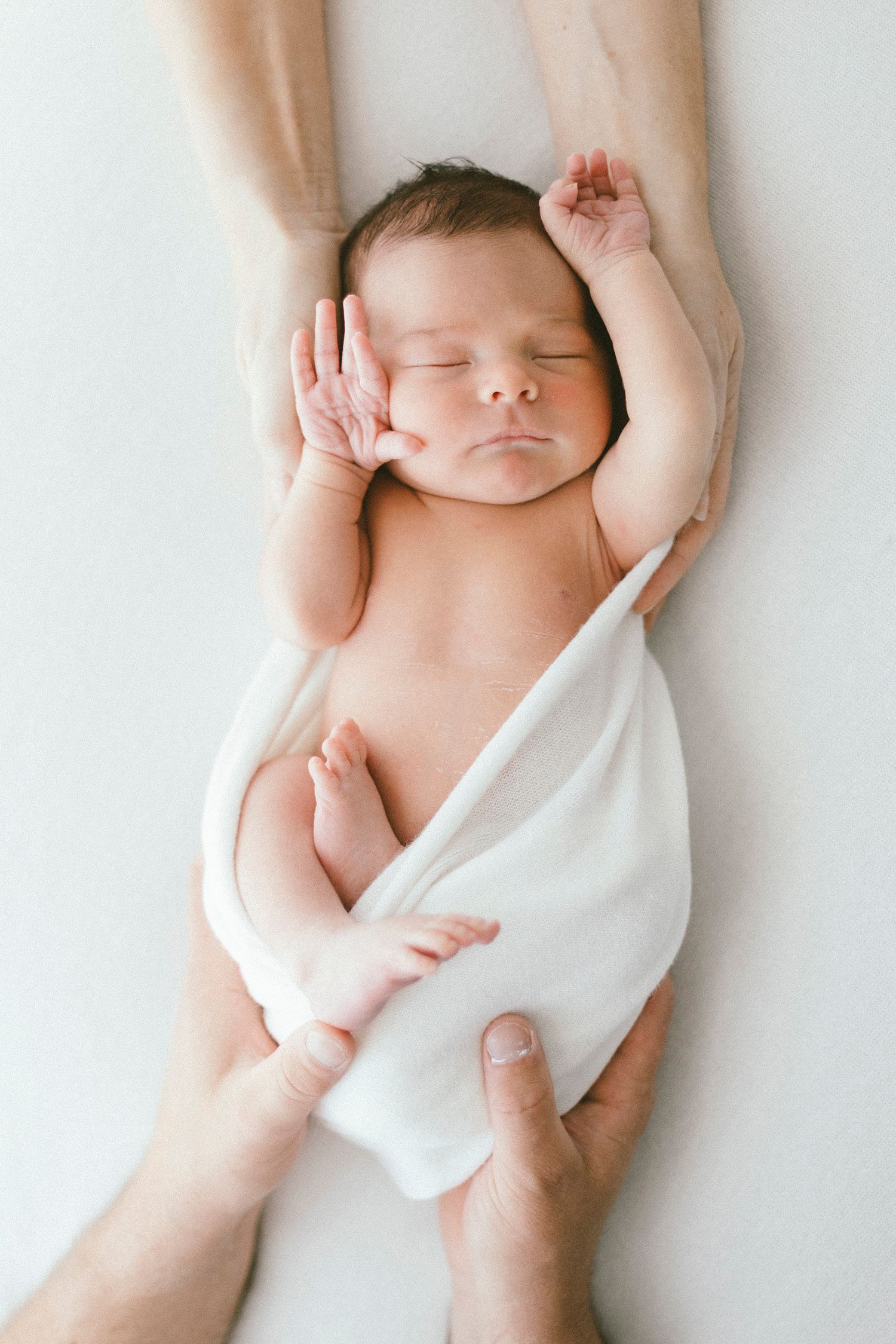Peaceful sleeping newborn wrapped in white fabric being held by caring hands.