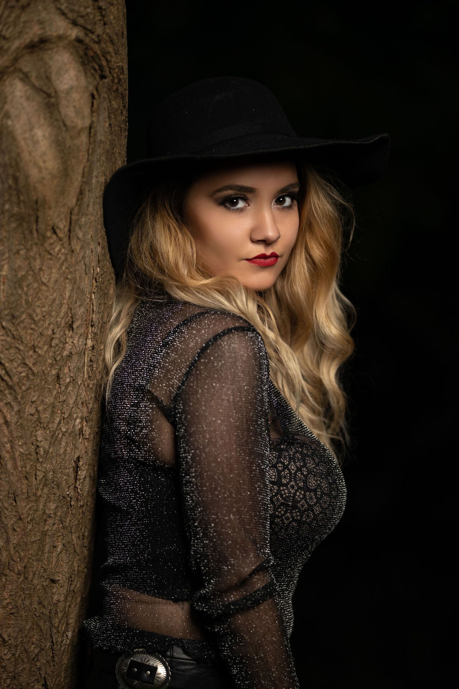 A dramatic portrait against a tree showing someone in a black hat and sheer lace top.