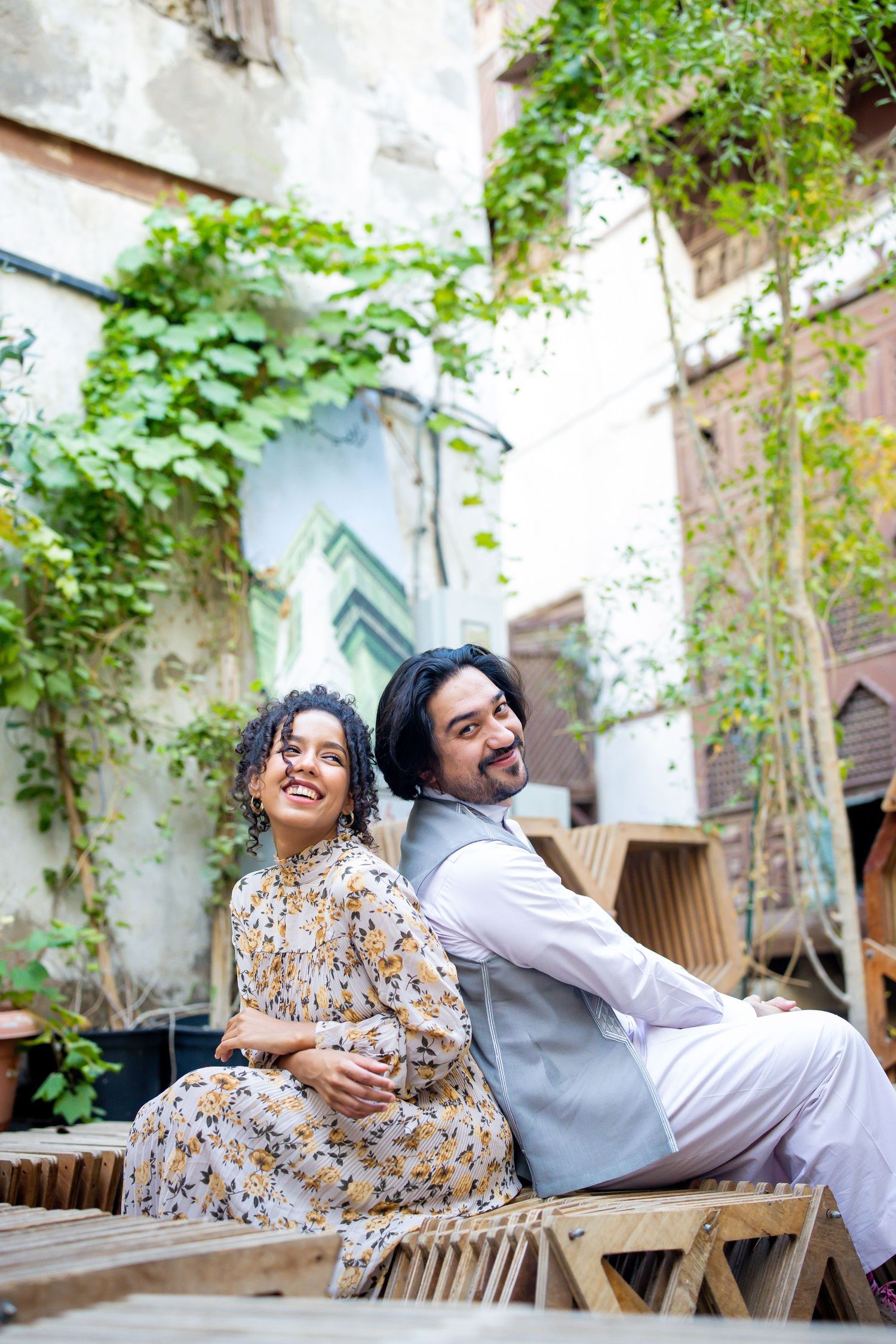 A couple shares joyful moment sitting back-to-back on wooden bench surrounded by greenery.