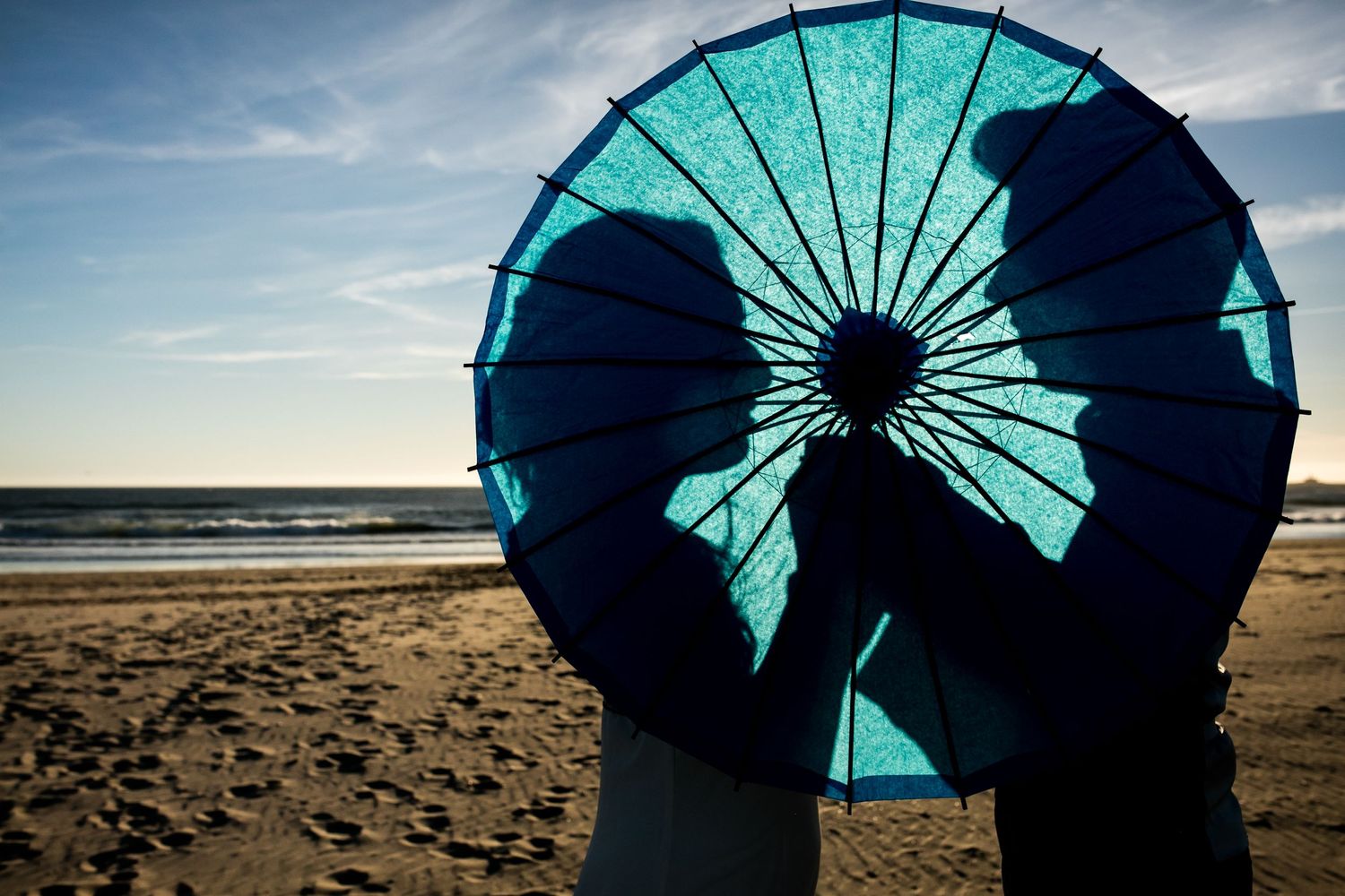 Silhouettes behind a turquoise parasol on a beach at sunset.
