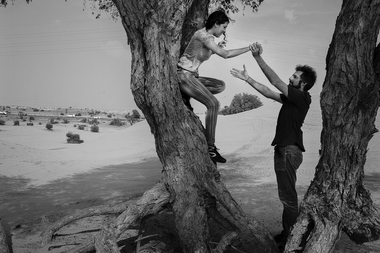 Two people playfully climb a tree in a desert landscape captured in black and white.