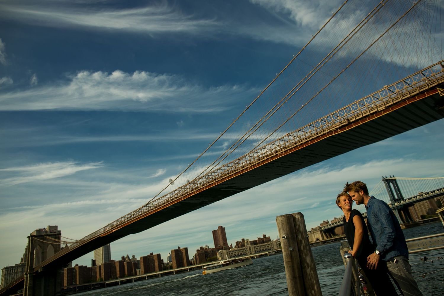 Couple embraces near waterfront with dramatic view of bridge and city skyline.
