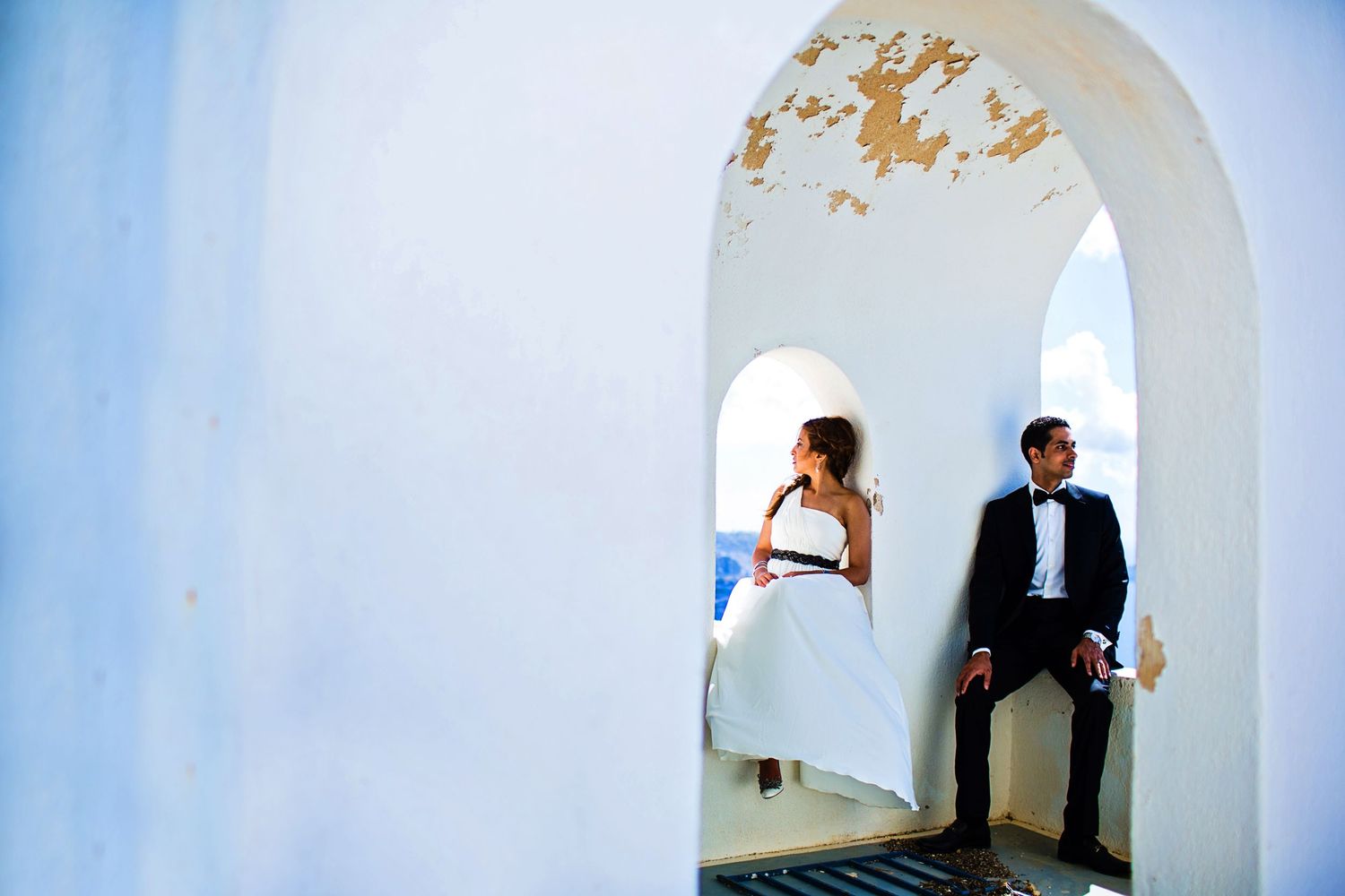 Artistic wedding portrait through a white archway against bright blue sky.